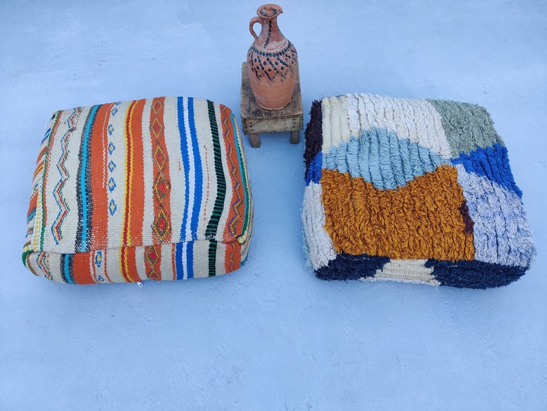 K&ouml;nnte beinhalten: Zwei bunte, gemusterte Bodenkissen, eines mit einem gestreiften Design und das andere mit einem Patchwork-Design, sind auf einer hellblauen Oberfl&auml;che abgebildet. Ein kleiner Holzhocker mit einer braunen Tonkrug darauf ist ebenfalls zu sehen.