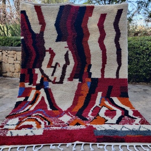 May include: A colorful Moroccan rug with a geometric pattern in shades of red, orange, blue, black, and white. The rug has a fringe border and is made of wool.