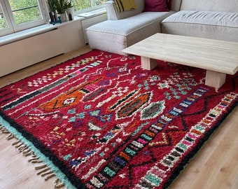 Könnte beinhalten: Ein roter, blauer und grüner Teppich mit Muster und Fransenrand. Der Teppich liegt auf einem Holzboden vor einem weißen Sofa und einem Holz-Couchtisch.