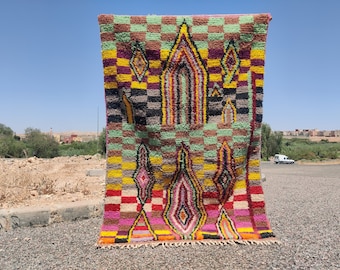 May include: A colorful, handwoven rug with a geometric design. The rug features a checkerboard pattern in green, brown, and yellow, with diamond and arch-shaped motifs in shades of pink, purple, and orange. The rug is displayed outdoors.