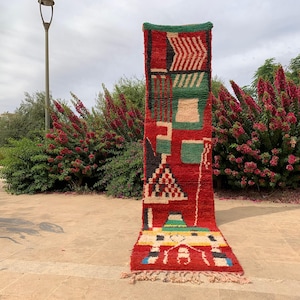 May include: A long, rectangular, hand-woven rug with a vibrant geometric design. The rug features a red base with green, black, white, and yellow accents. The design includes shapes like triangles, squares, and zigzags. The rug has a fringed edge.