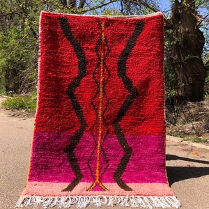 Puede incluir: Una alfombra marroquí roja, rosa y naranja con un patrón geométrico negro. La alfombra tiene flecos blancos en la parte inferior.
