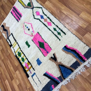 Peut inclure: Un tapis blanc avec un motif g&eacute;om&eacute;trique dans des tons de rose, bleu, vert, noir et jaune. Le tapis a une bordure frang&eacute;e.
