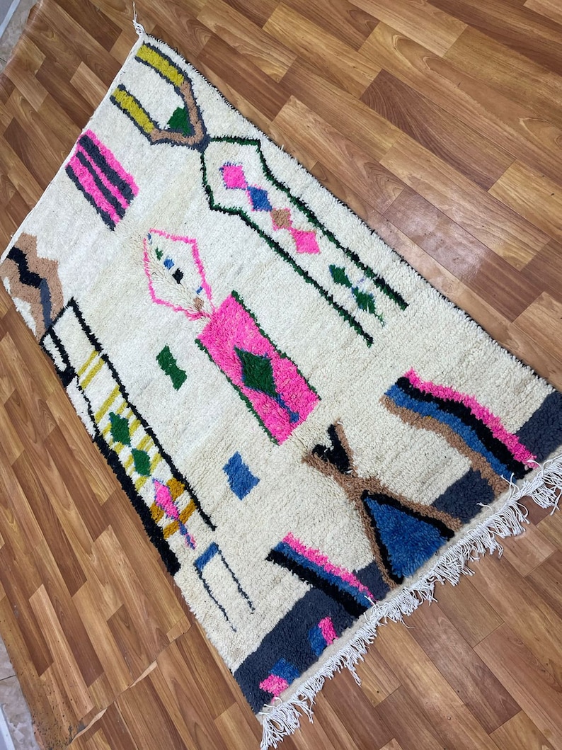 Peut inclure: Un tapis blanc avec un motif g&eacute;om&eacute;trique dans des tons de rose, bleu, vert, noir et jaune. Le tapis a une bordure frang&eacute;e.