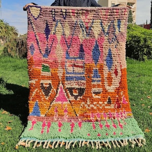 Puede incluir: Una alfombra marroquí colorida con un patrón geométrico en tonos de rosa, naranja, azul y verde. La alfombra tiene un borde de flecos y está hecha de lana.