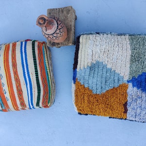 K&ouml;nnte beinhalten: Zwei farbenfrohe gewebte Poufs, einer mit einem Streifenmuster in Orange, Wei&szlig; und Blau, der andere mit einem Patchwork-Design in Blau, Wei&szlig; und Braun. Ein kleiner Holzhocker mit einem Tontopf steht im Hintergrund.