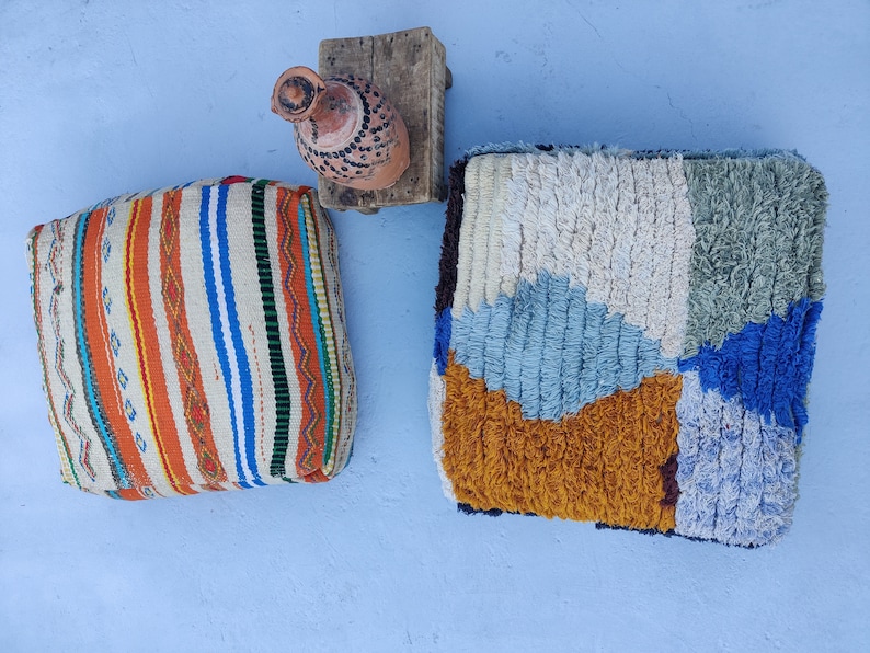 K&ouml;nnte beinhalten: Zwei farbenfrohe gewebte Poufs, einer mit einem Streifenmuster in Orange, Wei&szlig; und Blau, der andere mit einem Patchwork-Design in Blau, Wei&szlig; und Braun. Ein kleiner Holzhocker mit einem Tontopf steht im Hintergrund.