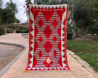 May include: A red and white Moroccan rug with a geometric pattern. The rug has a fringe border and is hanging on a wall.