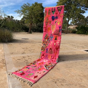 May include: A bright pink runner rug with a geometric pattern and fringing. The rug features a variety of colourful shapes and designs, including blue, green, and yellow accents. The rug is displayed outdoors on a stone surface.