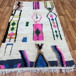 Peut inclure: Un tapis marocain blanc avec un motif g&eacute;om&eacute;trique dans des tons de rose, bleu, vert et marron. Le tapis a une bordure frang&eacute;e.