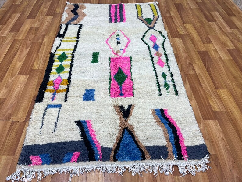 Peut inclure: Un tapis marocain blanc avec un motif g&eacute;om&eacute;trique dans des tons de rose, bleu, vert et marron. Le tapis a une bordure frang&eacute;e.