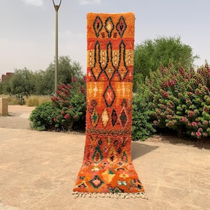May include: A vertical, rectangular rug with a vibrant orange and yellow color scheme. The rug features geometric patterns in black, green, and white, including diamond and abstract shapes. The rug is displayed outdoors, showcasing its intricate design and texture.