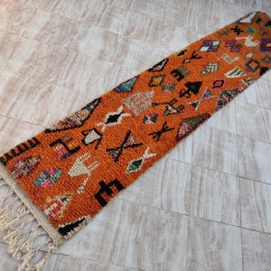 May include: A long, orange runner rug with a geometric pattern in black, white, and blue. The rug has a fringe on one end.