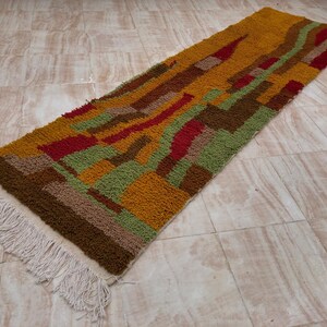 May include: A rectangular, colorful rug with a geometric pattern. The rug features a mix of colors including mustard yellow, red, green, and brown. The rug has a fringe on one end. The rug is approximately 60 inches long.