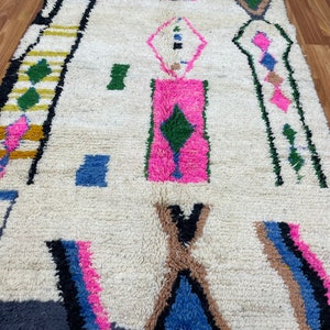 Peut inclure: Un tapis marocain blanc avec un motif g&eacute;om&eacute;trique dans des tons de rose, bleu, vert, marron, noir et jaune. Le tapis a une texture tiss&eacute;e &agrave; la main et une bordure &agrave; franges.