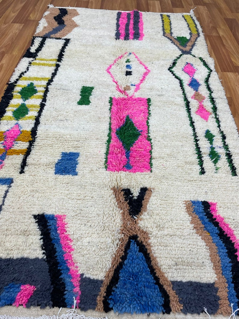 Peut inclure: Un tapis marocain blanc avec un motif g&eacute;om&eacute;trique dans des tons de rose, bleu, vert, marron, noir et jaune. Le tapis a une texture tiss&eacute;e &agrave; la main et une bordure &agrave; franges.