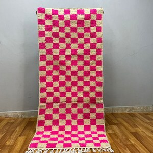 Puede incluir: Una alfombra a cuadros rosa y crema con flecos en el borde inferior. La alfombra está hecha de un material suave y tejido.