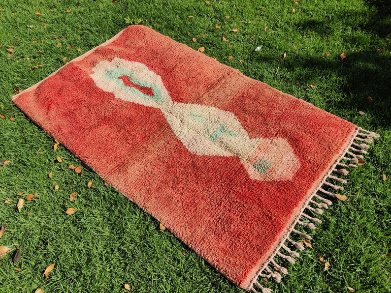 Puede incluir: Una alfombra roja marroqu&iacute; con un patr&oacute;n geom&eacute;trico blanco y verde. La alfombra tiene flecos en los bordes.