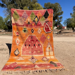 Puede incluir: Una alfombra marroquí colorida con un patrón geométrico en tonos de rojo, naranja, verde y negro. La alfombra está tejida con una variedad de texturas y tiene un borde de flecos.