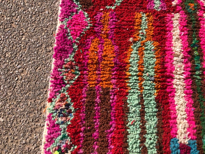 Puede incluir: Primer plano de una alfombra marroqu&iacute; colorida con rayas de color rosa, rojo, naranja, verde, blanco y marr&oacute;n. La alfombra est&aacute; hecha de lana y tiene una textura suave y esponjosa.