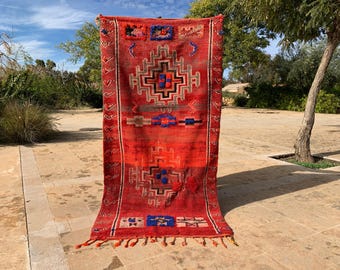 May include: A vibrant red and orange rug with geometric patterns in blue, black, and beige. The rug features a central diamond-like design and decorative borders. Tassels adorn the bottom edge. This handmade rug is suitable for home decor.