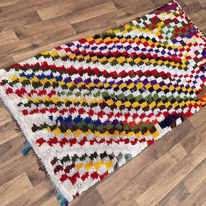 May include: A colourful Moroccan rug with a geometric pattern in shades of red, white, yellow, green, blue, and purple. The rug has a hand-woven texture and is made of wool.