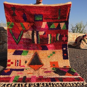 May include: A colorful Moroccan rug with geometric patterns in shades of red, pink, green, blue, and black. The rug has a fringe border and is made of wool.
