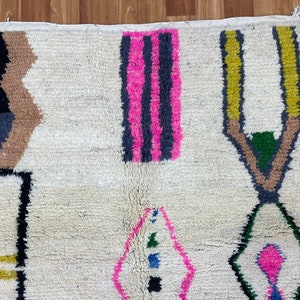 Peut inclure: Un tapis blanc avec un motif g&eacute;om&eacute;trique en noir, marron, rose, jaune et vert. Le tapis a une bordure frang&eacute;e.