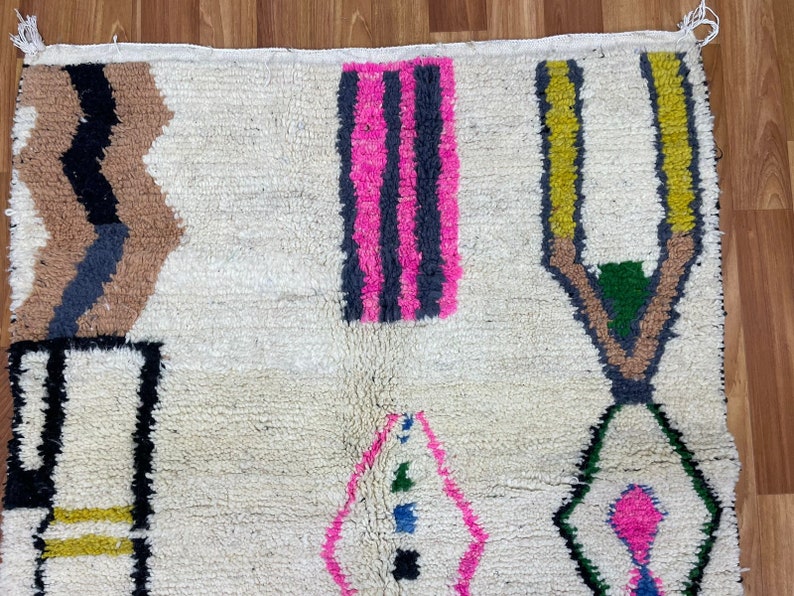 Peut inclure: Un tapis blanc avec un motif g&eacute;om&eacute;trique en noir, marron, rose, jaune et vert. Le tapis a une bordure frang&eacute;e.