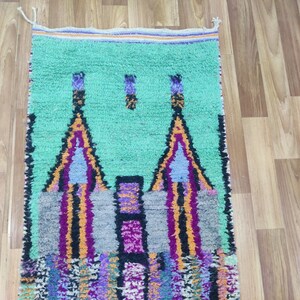 May include: A hand-woven Moroccan rug with a green background and a colourful geometric design featuring two towers. The rug has a fringe border.