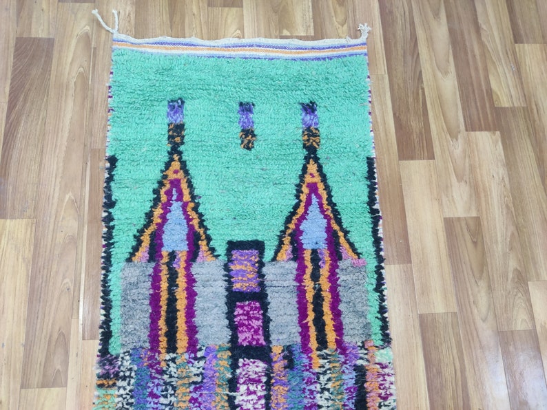 May include: A hand-woven Moroccan rug with a green background and a colourful geometric design featuring two towers. The rug has a fringe border.