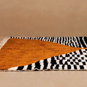 May include: A black, white, and mustard yellow patterned rug with fringe. The rug has a geometric design with stripes, squares, and a large triangle shape.