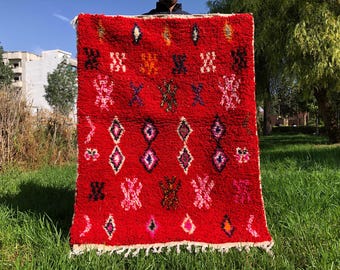 May include: A red Moroccan rug with a geometric pattern featuring diamonds, stars, and other abstract shapes. The rug has a fringe border and is made of wool.