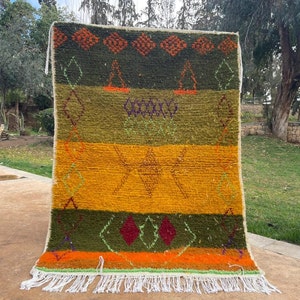 May include: A handwoven Moroccan rug with a geometric pattern in shades of green, orange, yellow, and red. The rug has a fringe border and is hanging on a wall.