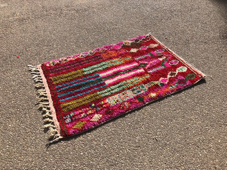 Puede incluir: Una alfombra marroqu&iacute; colorida con un patr&oacute;n geom&eacute;trico en tonos de rosa, rojo, verde, azul y marr&oacute;n. La alfombra tiene un borde de flecos y est&aacute; hecha de lana.
