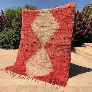 May include: A vibrant, hand-woven rug with a geometric design. The rug features a bold red background with a large, diamond-shaped pattern in a natural beige color. The rug has a fringed edge.