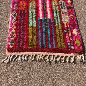 Puede incluir: Una alfombra marroqu&iacute; colorida con un patr&oacute;n de rayas rosa, rojo, verde, azul y marr&oacute;n. La alfombra tiene un borde de flecos y est&aacute; hecha de lana.