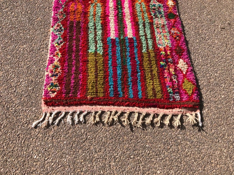 Puede incluir: Una alfombra marroqu&iacute; colorida con un patr&oacute;n de rayas rosa, rojo, verde, azul y marr&oacute;n. La alfombra tiene un borde de flecos y est&aacute; hecha de lana.