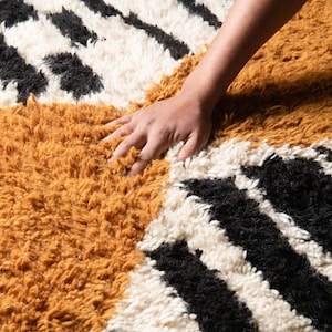 May include: A hand rests on a fluffy rug with a geometric pattern of black, white, and mustard yellow stripes.