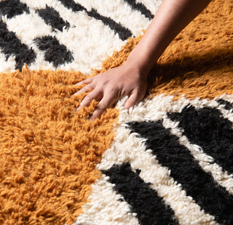 May include: A hand rests on a fluffy rug with a geometric pattern of black, white, and mustard yellow stripes.