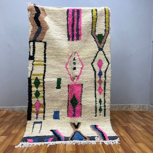 Peut inclure: Un tapis blanc avec un motif g&eacute;om&eacute;trique en rose, vert, noir et marron. Le tapis a une bordure &agrave; franges.