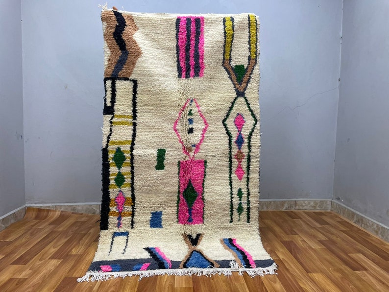 Peut inclure: Un tapis blanc avec un motif g&eacute;om&eacute;trique en rose, vert, noir et marron. Le tapis a une bordure &agrave; franges.