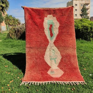 Puede incluir: Una alfombra roja marroqu&iacute; con un dise&ntilde;o geom&eacute;trico blanco y verde. La alfombra tiene un borde de flecos y est&aacute; colgada de un tendedero.