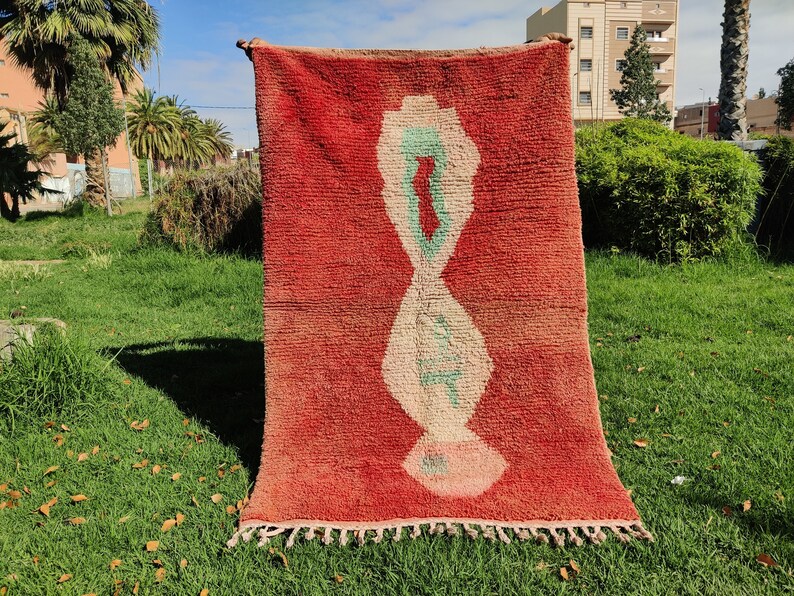 Puede incluir: Una alfombra roja marroqu&iacute; con un dise&ntilde;o geom&eacute;trico blanco y verde. La alfombra tiene un borde de flecos y est&aacute; colgada de un tendedero.