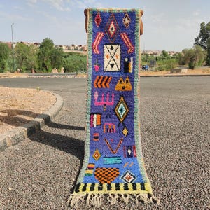 Puede incluir: Una alfombra larga y estrecha con fondo azul y patrones geométricos coloridos. La alfombra tiene flecos en ambos extremos.