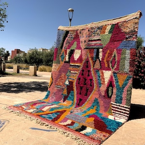 May include: A vibrant, hand-woven rug with a geometric design. The rug features a mix of colors, including pink, blue, orange, and green. The rug is displayed outdoors, showcasing its intricate patterns and textures. The rug has a fringed edge.