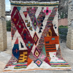 May include: A colorful Moroccan rug with a geometric pattern in shades of red, orange, blue, and green. The rug has a white background and is woven with a variety of textures.
