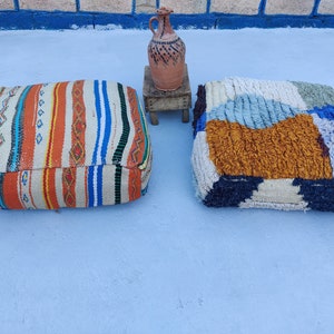K&ouml;nnte beinhalten: Zwei bunte, gemusterte Bodenkissen, eines mit einem geometrischen Muster in Blaut&ouml;nen, Wei&szlig; und Braun, das andere mit einem Streifenmuster in Orange, Blau und Wei&szlig;. Die Kissen liegen auf einer hellblauen Oberfl&auml;che.