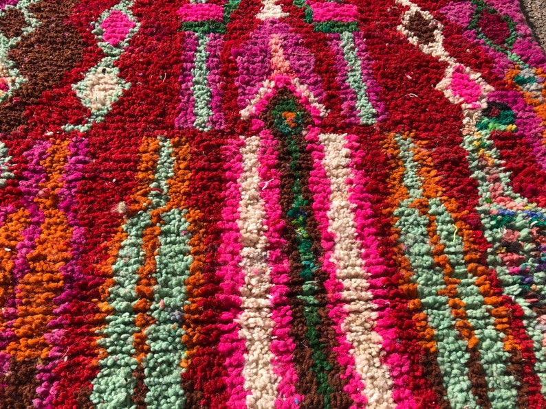 Puede incluir: Primer plano de una alfombra marroqu&iacute; colorida con un patr&oacute;n geom&eacute;trico. La alfombra est&aacute; hecha de lana y tiene una textura suave y esponjosa. Los colores son brillantes y vibrantes, incluyendo rojo, rosa, naranja, verde y blanco.