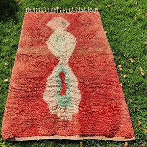 Puede incluir: Una alfombra roja marroqu&iacute; con un patr&oacute;n geom&eacute;trico blanco y verde. La alfombra tiene un borde de flecos.